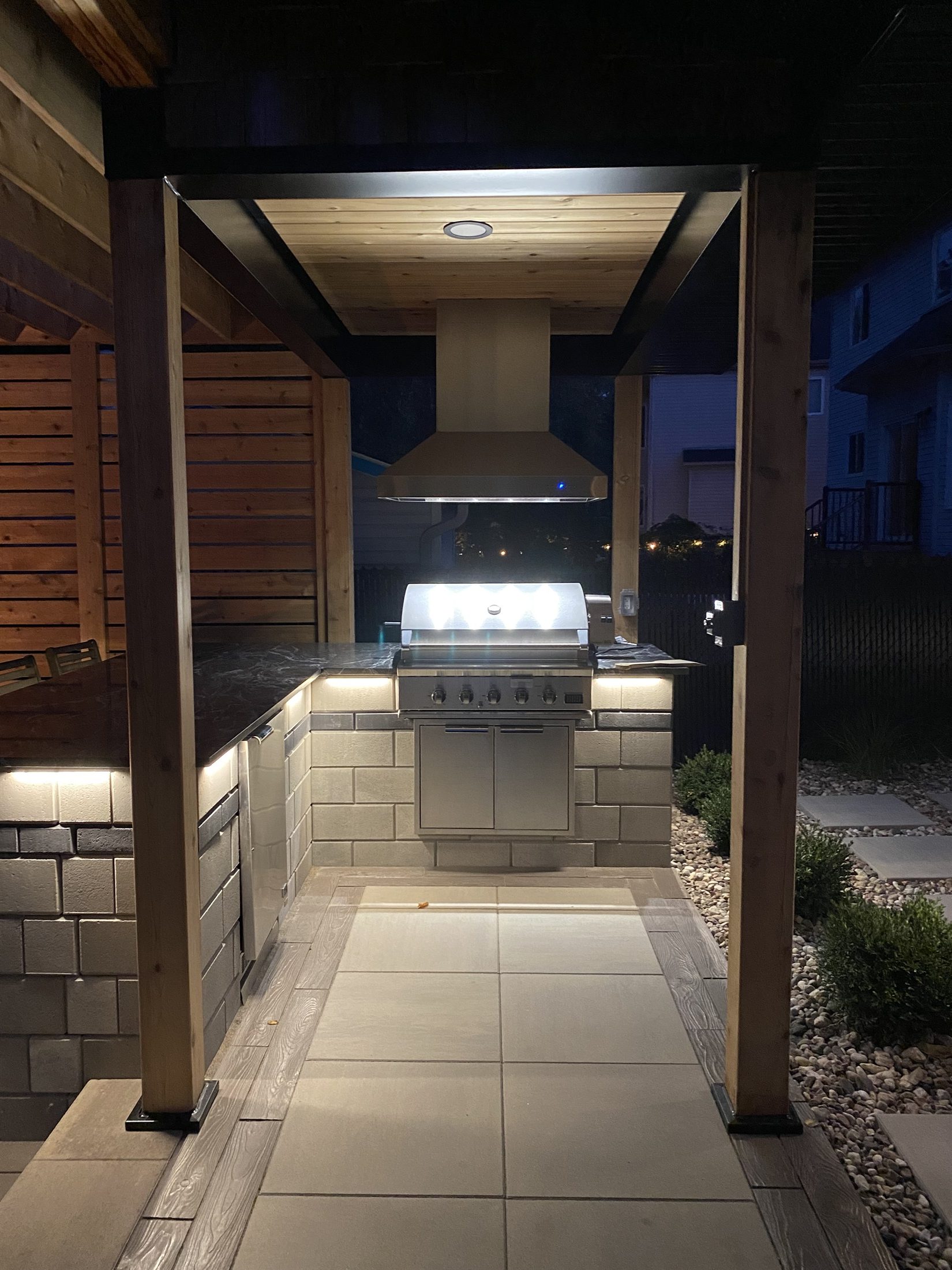 Outdoor kitchen at night with bbq, hood vent, fridge and stonework.